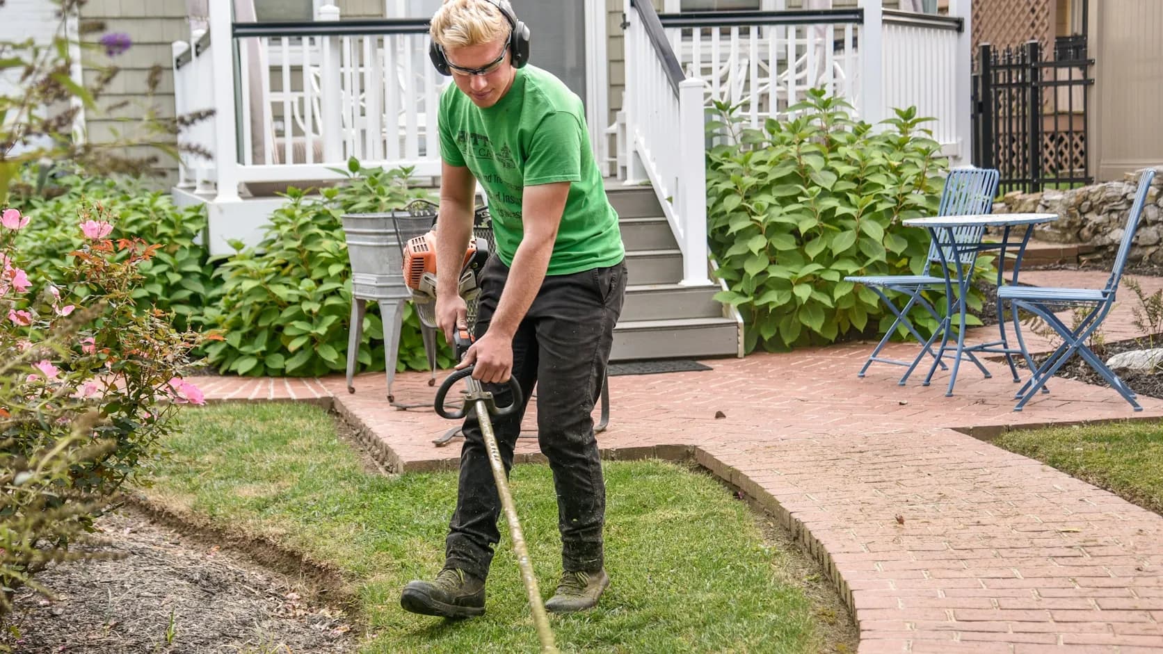 A lawn care worker in a green shirt and ear protection running a string trimmer to edge a residential lawn beside a house — a crew member actively at work.